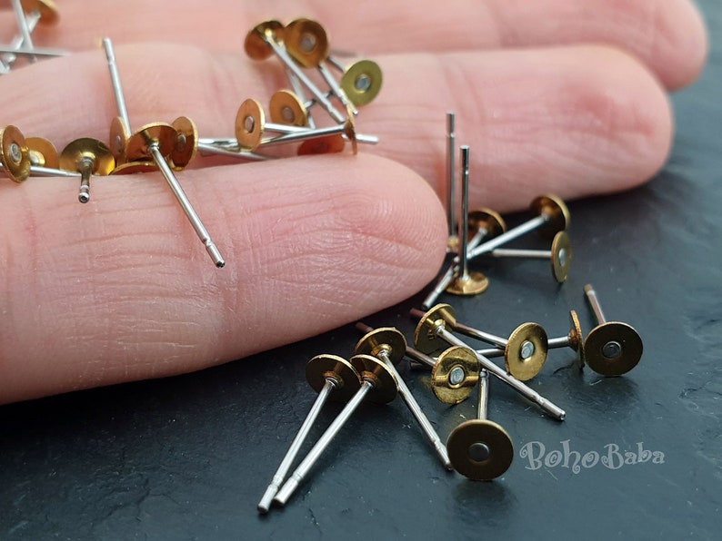 Puede incluir: Primer plano de un mont&oacute;n de postes de pendientes de color dorado con tallos plateados. Los postes de pendientes est&aacute;n esparcidos sobre una superficie oscura.