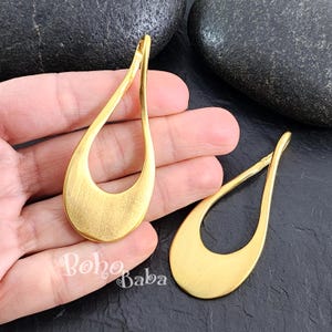 May include: Two gold-toned metal teardrop-shaped earring blanks with a large open circle in the center. The blanks are shown on a hand.