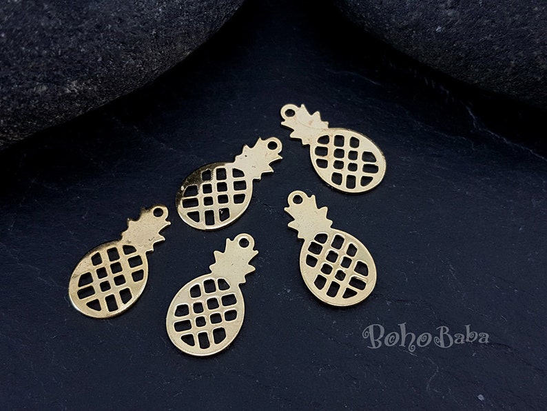 May include: Five gold metal pineapple charms with a cutout design. The charms are on a black background.