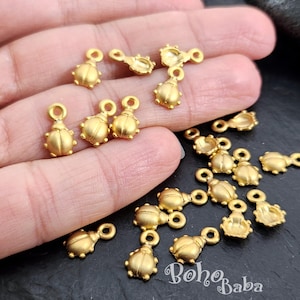 Peut inclure: Gros plan sur une main tenant une variété de breloques scarabées en métal doré. Les breloques ont une surface texturée et sont parfaites pour ajouter une touche de fantaisie à vos projets de bijoux.