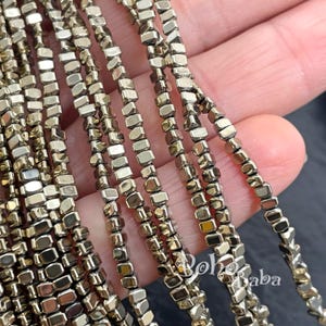 May include: Close-up of multiple strands of metallic, rectangular beads. The beads have a gold and silver color, and are strung together to form a necklace or bracelet. The beads are small and have a faceted appearance.