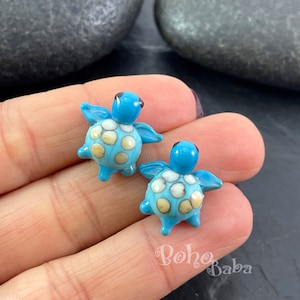Op de afbeelding: Een paar blauwe glazen schildpad oorbellen met witte en gele vlekken. De oorbellen zijn klein en delicaat, en ze zouden een geweldig cadeau zijn voor iemand die van schildpadden houdt.