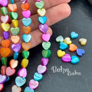 Op de afbeelding: Een streng iriserende hartvormige kralen in een regenboog van kleuren. De kralen zijn gemaakt van parelmoer en geregen aan een doorzichtig elastisch koord. De kralen zijn ongeveer 1,25 cm in diameter. De kralen worden getoond op een hand en op een donkergrijs oppervlak. De tekst "boho baba" is zichtbaar in de afbeelding.