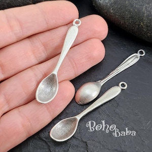 May include: Two silver spoon charms with a loop for hanging. The spoons are slightly tarnished and have a vintage look.