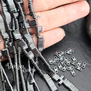 Peut inclure: Gros plan sur un brin de perles d'hématite noire, avec un tas de petites perles d'espacement argentées en arrière-plan. Les perles sont rectangulaires et ont une finition lisse et polie.