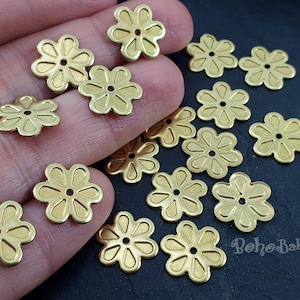 May include: A collection of gold metal flower-shaped embellishments with a hole in the center. The embellishments are scattered on a hand and a dark surface.
