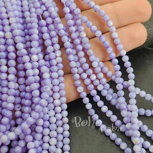 Puede incluir: Una hebra de cuentas de concha de color lavanda. Las cuentas son redondas y tienen un ligero brillo iridiscente. Las cuentas están enhebradas en un hilo transparente.