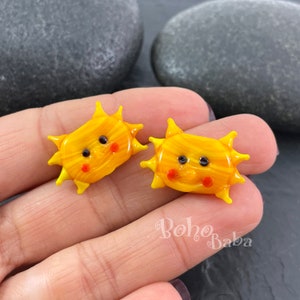 May include: Two yellow glass sun earrings with a smiling face, black eyes, and red cheeks. The earrings are on a hand.