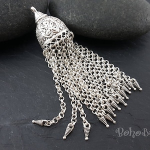 May include: Silver tassel pendant with a detailed, ornate top and a long chain of silver links. The tassel is made of multiple strands of chain with small, pointed tips.
