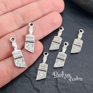 May include: Six silver paintbrush charms, each with a detailed brush head and handle. The charms are displayed on a hand against a dark background.
