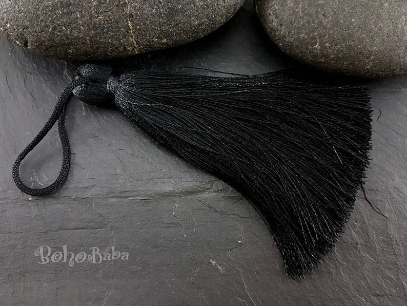 K&ouml;nnte beinhalten: Ein schwarzer Quastenanh&auml;nger mit einer Schlaufe zum Aufh&auml;ngen. Der Quastenanh&auml;nger besteht aus vielen Str&auml;ngen schwarzem Faden. Das Wort "Boho Baba" ist in wei&szlig;er Schrift auf der Oberfl&auml;che darunter gedruckt.