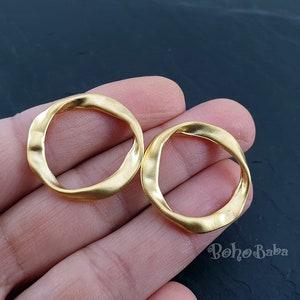 May include: Two gold-coloured, open-circle earrings with a textured, wavy design. The earrings have a matte finish and a simple, elegant style. The earrings are likely made of metal and are suitable for everyday wear.