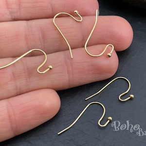 Peut inclure: Quatre crochets d'oreille dorés avec une petite boule à l'extrémité de chaque crochet. Les crochets sont présentés sur une main.