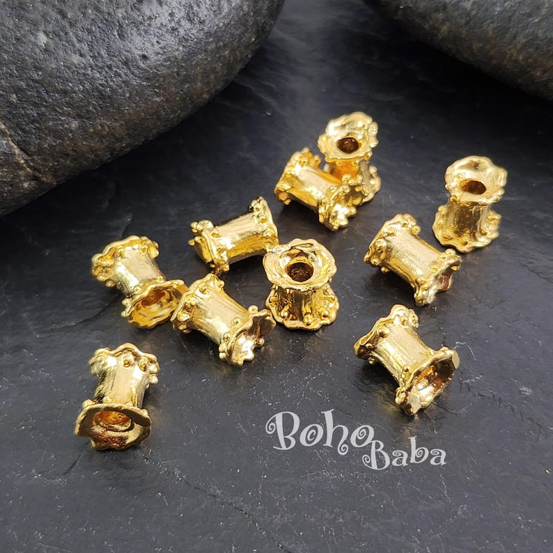 May include: A group of 10 gold-colored metal beads with a decorative, ornate design. The beads are arranged on a dark gray surface.