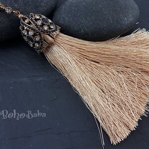 May include: A decorative tassel featuring a bronze-toned metal cap embellished with black and clear crystals. The tassel is a light tan colour, composed of numerous fine strands. The text "BohoBaba" is visible in the image.