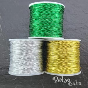 May include: Three spools of metallic thread in green, silver, and gold. The spools are stacked, with the green spool on top. The silver and gold spools are side by side. The thread is tightly wound on white plastic spools.