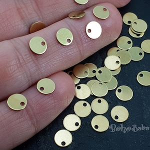 Puede incluir: Primer plano de una mano sosteniendo un montón de pequeños discos metálicos redondos de color dorado con un agujero en el centro. Los discos están esparcidos sobre una superficie negra.