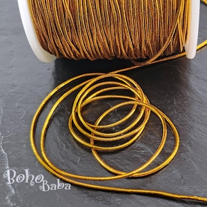May include: A spool of gold elastic cord. The cord is wound around a white spool and is lying on a dark gray surface.