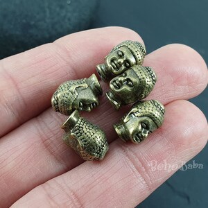 May include: Five antique gold-toned metal Buddha head beads. The beads are detailed with a textured surface and have a slightly worn look.