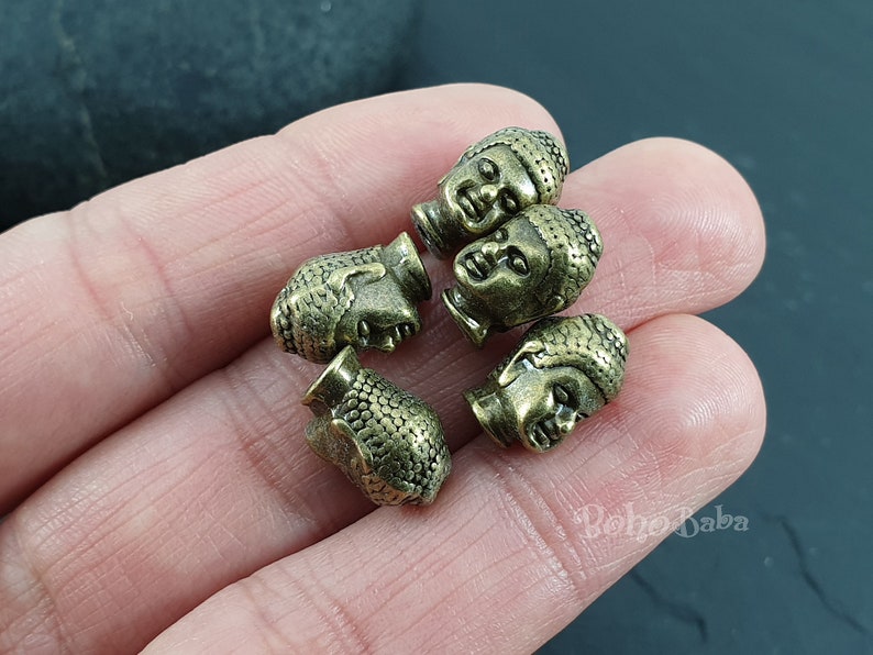 May include: Five antique gold-toned metal Buddha head beads. The beads are detailed with a textured surface and have a slightly worn look.