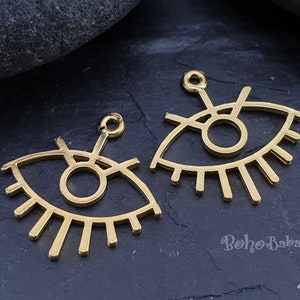 Puede incluir: Dos abalorios de metal de color dorado en forma de ojo con un c&iacute;rculo en el centro y pesta&ntilde;as alrededor. Los abalorios tienen un bucle en la parte superior para sujetarlos a joyas.