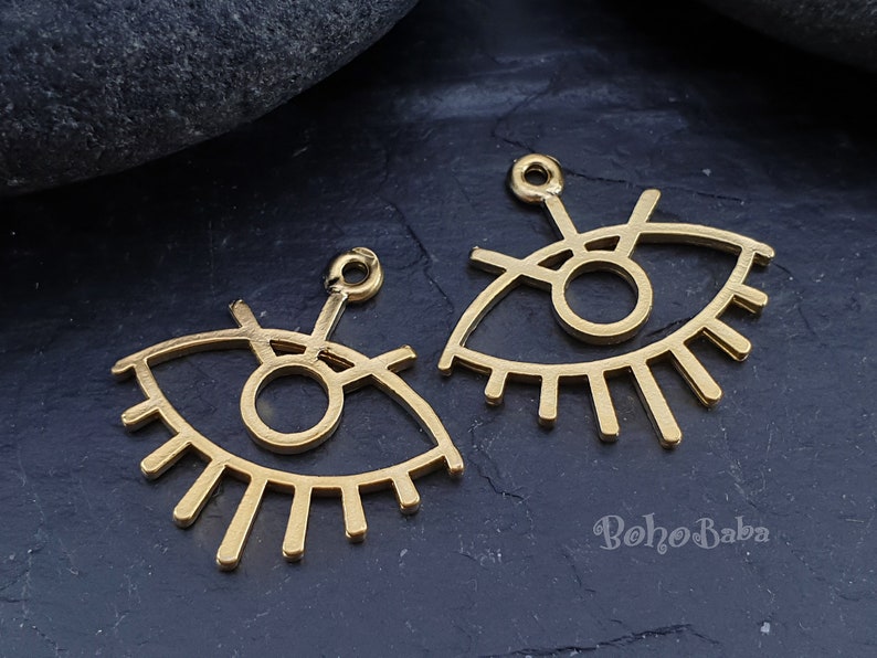 Puede incluir: Dos abalorios de metal de color dorado en forma de ojo con un c&iacute;rculo en el centro y pesta&ntilde;as alrededor. Los abalorios tienen un bucle en la parte superior para sujetarlos a joyas.