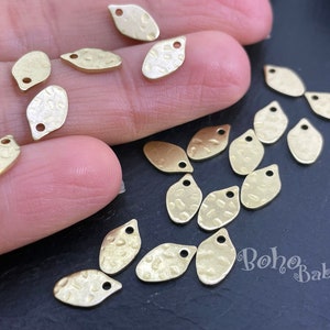 Puede incluir: Una colección de 17 abalorios de metal de color dorado en forma de hoja con textura martillada. Los abalorios tienen un agujero en la parte superior para sujetarlos a joyas u otras manualidades.