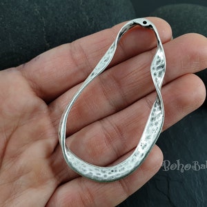 May include: Silver-toned metal pendant with a twisted, hammered, and oval shape. The pendant has a hole at the top for attaching a chain or cord.