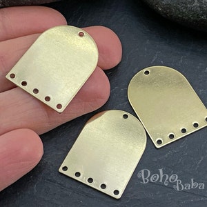 Op de afbeelding: Drie goudkleurige metalen boogvormige bedels met elk vijf gaten. De bedels liggen op een hand en een donker oppervlak.