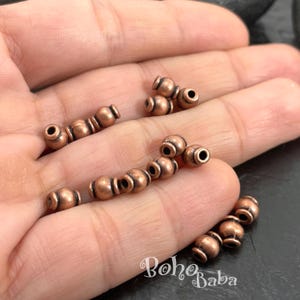 Peut inclure: Un ensemble de petites perles cylindriques couleur cuivre avec une finition métallique. Chaque perle présente une section centrale arrondie flanquée de deux anneaux plus fins. Les perles sont éparpillées, affichant une teinte brun-rouge.