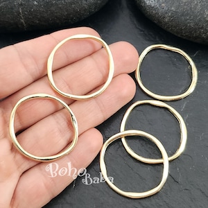 Puede incluir: Varios anillos abiertos dorados de forma irregular. Los anillos son lisos y brillantes, con un borde ligeramente irregular. Los anillos se muestran sobre una superficie oscura. El logotipo de Boho Baba es visible.