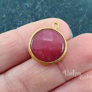 Puede incluir: Un colgante dorado con una piedra redonda de cabujón rosa. El colgante tiene un diseño simple y elegante y sería una hermosa adición a cualquier colección de joyas.