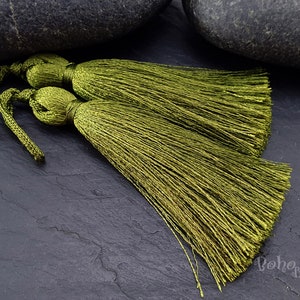 Op de afbeelding: Twee olijfgroene zijden kwastjes met een lange franje. De kwastjes zijn aan elkaar gebonden met een dun koord.