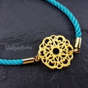 May include: A turquoise cord bracelet with a gold filigree charm. The charm is a circular design with a center hole.