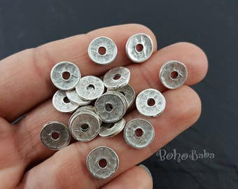 May include: A collection of silver-toned metal spacer beads with a round, flat shape and a hole in the center. The beads are arranged in a pile on a hand.