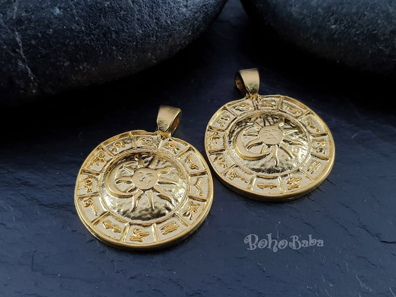 May include: Two gold-toned pendants with a sun and moon design and zodiac symbols around the edge. The pendants are on a dark gray background.