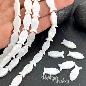 Może przedstawiać: Sznury białych koralików w kształcie ryb. Koraliki wykonane są z perłowego materiału i są nawleczone. Kilka luźnych koralików leży na ciemnej powierzchni. Tekst "Boho Baba" jest widoczny w prawym dolnym rogu.