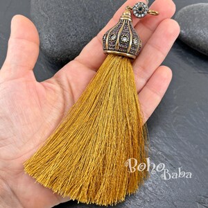 May include: A decorative tassel featuring a metallic gold-toned cap with an ornate, dome-shaped design. The cap is embellished with small, clear, sparkling stones and a dark, textured finish. A long, dense fringe of shimmering golden-yellow thread hangs below.