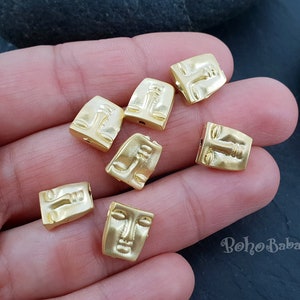 Könnte beinhalten: Nahaufnahme von sieben goldfarbenen Metallperlen mit Gesichtsmotiven. Die Perlen sind rechteckig und haben ein erhabenes Gesichtsdesign. Die Perlen sind auf einer Hand angeordnet, wobei die Finger der Hand gespreizt sind.