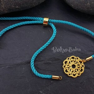 May include: Teal blue cord bracelet with a gold adjustable slider and a gold filigree charm. The charm is a circular design with a mandala pattern. The bracelet is on a dark gray background.