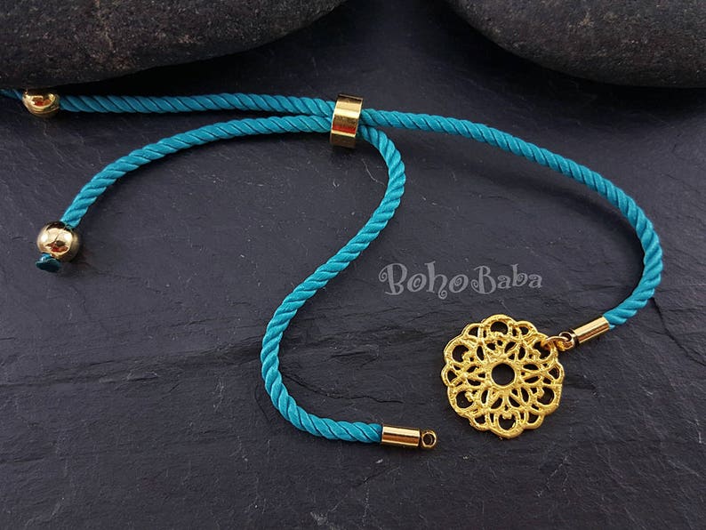 May include: Teal blue cord bracelet with a gold adjustable slider and a gold filigree charm. The charm is a circular design with a mandala pattern. The bracelet is on a dark gray background.