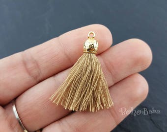 May include: A gold tassel with a hammered metal top and a light brown tassel. The tassel is held in a hand.