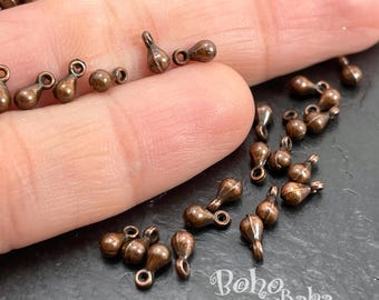 May include: A collection of small, copper-colored metal teardrop-shaped charms, each featuring a tiny circular loop at the top for attachment. The charms have a smooth, metallic finish and are scattered across a dark, textured surface.