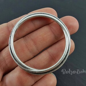 Può includere: Un anello in metallo argentato con una superficie liscia e arrotondata. L'anello è spesso e ha un bordo leggermente rialzato.