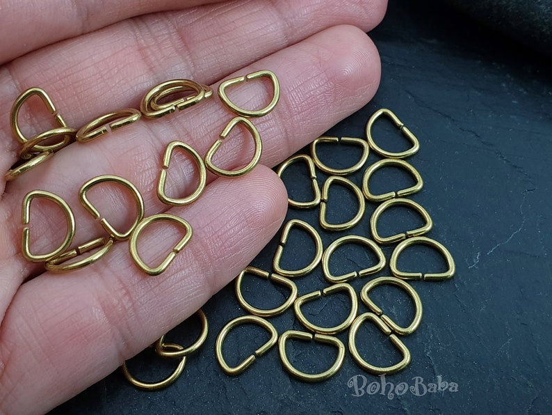 May include: A close-up of a hand holding a pile of gold-colored metal D-rings. The D-rings are all the same size and are arranged in a random pattern. There are about 30 D-rings in the pile.