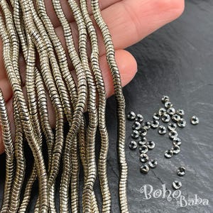 May include: Close-up of silver-colored, snake-like chain strands and small, round beads. The chains are held in a hand, with a dark background. The beads are scattered on the surface. The image is well-lit.