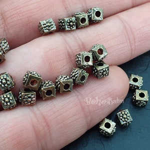 May include: A close-up of a collection of small, square, metal beads with a textured surface. The beads are a bronze color and have a hole in the center. The beads are scattered on a hand.