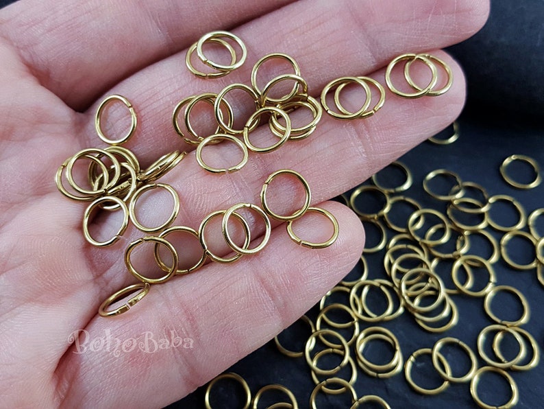 Peut inclure: Un grand nombre d'anneaux de saut dor&eacute;s, un type de composant de bijoux, sont &eacute;parpill&eacute;s sur une surface noire. Une main tient un petit groupe d'anneaux de saut.