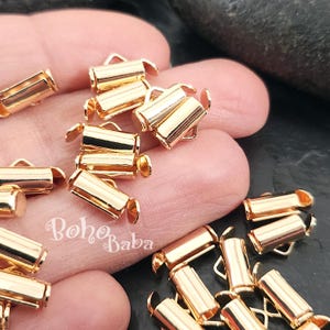 Op de afbeelding: Een verzameling kleine, goudkleurige metalen krimpeindstukken. Deze cilindrische onderdelen hebben aan elk uiteinde een lus en zijn ontworpen voor het maken van sieraden. De afbeelding toont verschillende stukken verspreid over een oppervlak, met het "Boho Baba" logo.