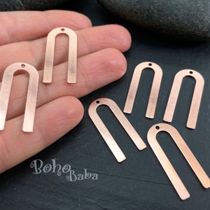 Puede incluir: Amuletos metálicos en forma de U de color cobre con un pequeño agujero en la parte superior para la fabricación de joyas. Los amuletos están dispuestos sobre una mano y una superficie oscura. La marca "Boho Baba" es visible.